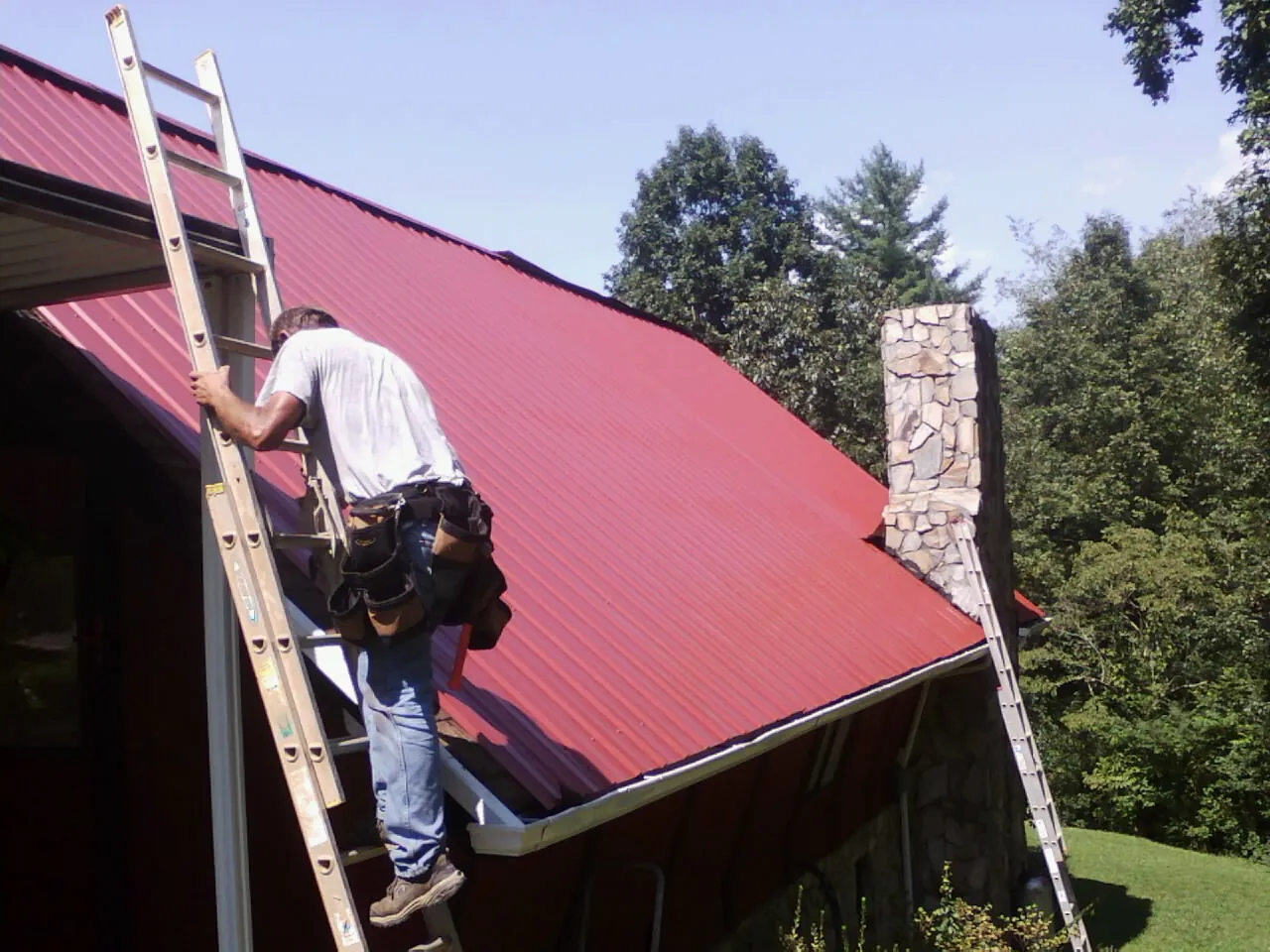 Roofer on a ladder beside a red metal roof and stone chimney — Soffit & Fascia Repair by Roofing Brothers in Hodgenville